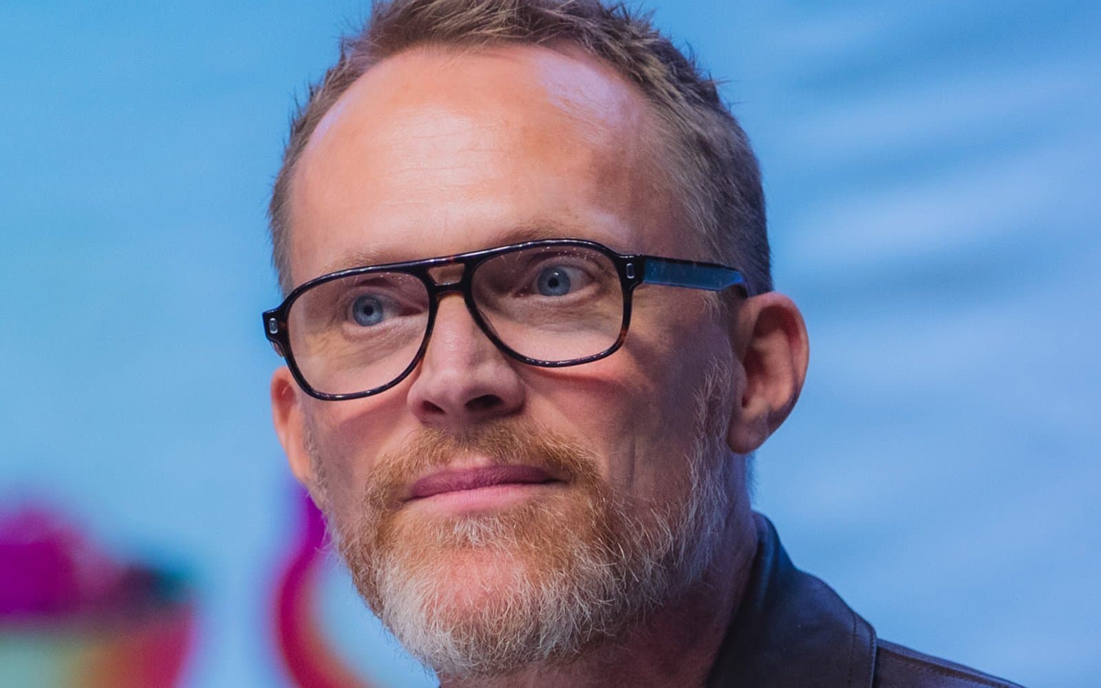Paul Bettany in dark-framed glasses and a brown leather jacket, smiling slightly on a convention stage against a bright blue backdrop.