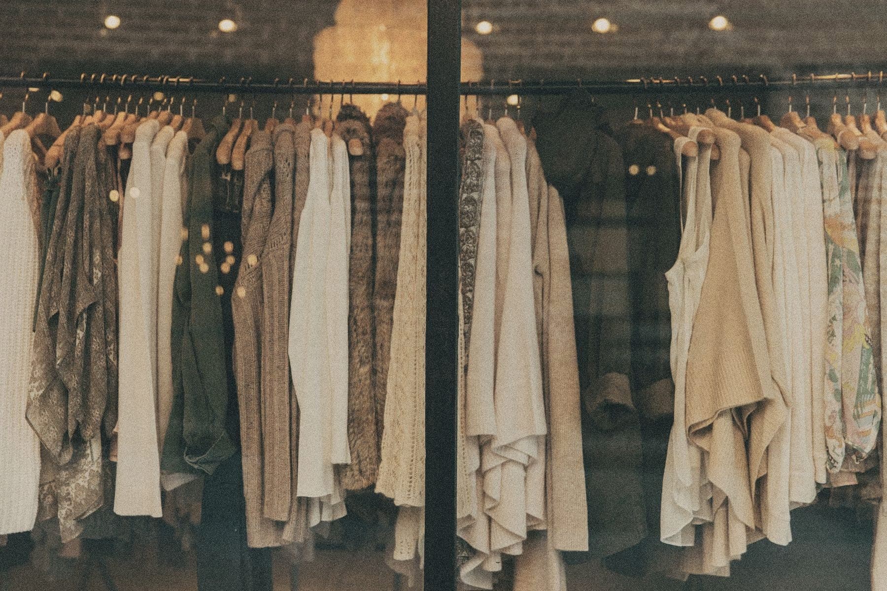 Racks of neutral-toned sweaters, knits, and shirts on wooden hangers, seen through a shop window.
