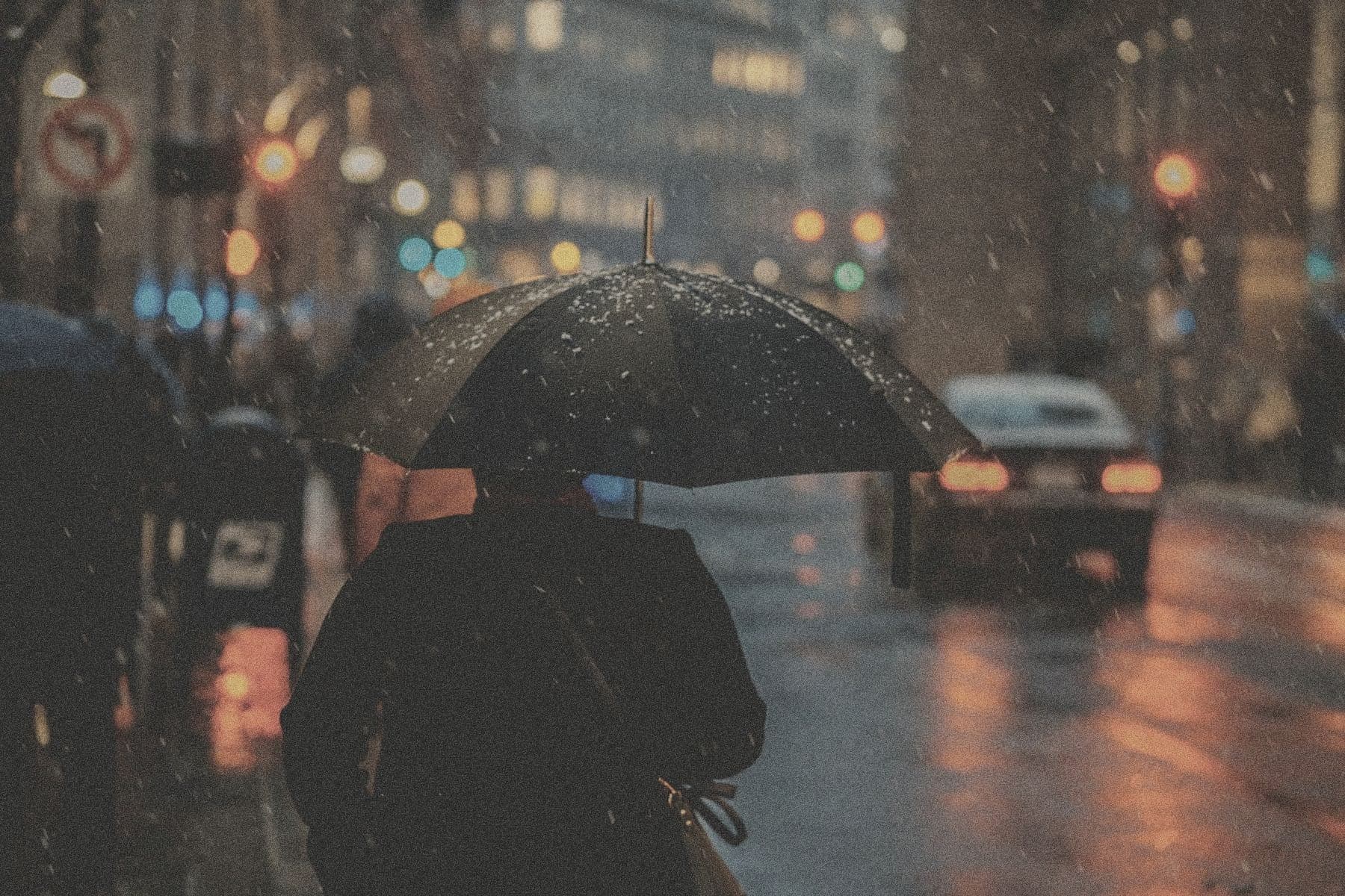 A person with an umbrella walking down a rainy city street at night, headlights and streetlights blurred in the background.